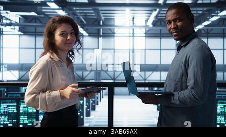 Portrait of happy system administrators in data center using devices, verifying firewall configurations. Jolly teamworking IT experts performing equipment vulnerability scans in server hub, camera B Stock Photo