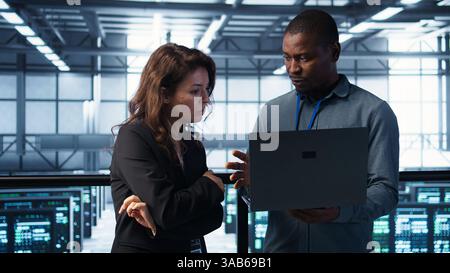 Developers in data center workspace coding on laptop, analyzing metrics, optimizing storage volumes. Teamworking IT professionals in server room using notebook, ensuring seamless operations, camera B Stock Photo