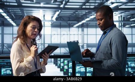 System administrators using notebook device in data center, managing server virtualization. IT specialists using laptop in server farm, sustaining long term infrastructure reliability, camera B Stock Photo