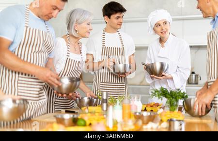 Female chef giving instructions to students standing around table in cooking class Stock Photo