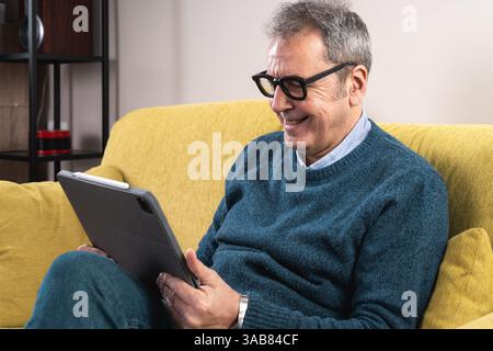 Happy senior man sitting on a couch using a tablet, smiling while browsing or watching digital content at home. Concept of modern technology, connecti Stock Photo