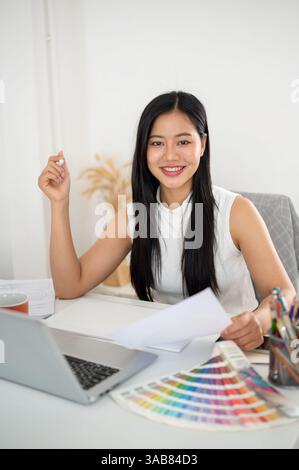 An asian woman is smiling at camera while holding the documents about design color on her working desk. Interior Designer or Graphic Designer working Stock Photo