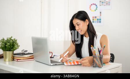 An asian woman is choosing the color from color charts or palettes while looking at the laptop screen. Interior Designer or Graphic Designer working i Stock Photo