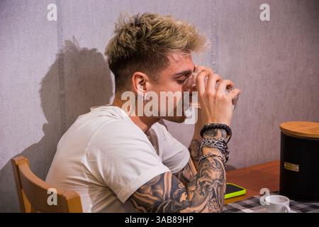 A young man with blond hair and blue eyes sits at a table, sipping a drink. Surrounded by a relaxed atmosphere, he showcases his tattoos and casual style while enjoying his moment. Stock Photo