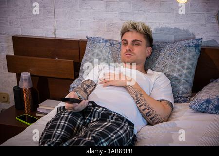 A young man with blond hair and blue eyes rests on a bed, dressed casually in a t-shirt and plaid pants. He focuses on the television, enjoying a calm evening at home. Stock Photo