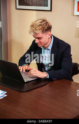 A handsome young man sits at a desk focused on his laptop. He has blond hair and blue eyes, dressed in a suit, clearly engaged in his work environment during daylight hours. Stock Photo