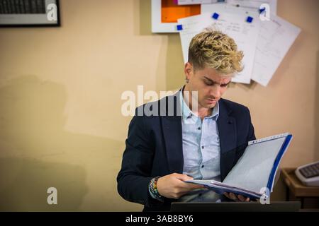 A young man with blond hair and blue eyes examines a folder filled with documents. He appears focused and engaged, seated at a desk in a contemporary office setting. Stock Photo