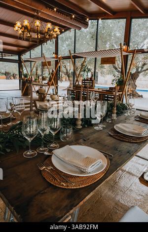 A rustic wooden banquet table with woven placemats, elegant glassware, striped napkins, and lush greenery in the center, all within a glass-walled ven Stock Photo