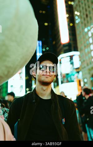 A young man wearing sunglasses and a cap enjoys the vibrant nightlife in Times Square. He stands confidently, surrounded by bright lights and bustling Stock Photo