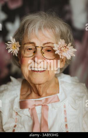 An elderly woman wearing glasses and flowers in her hair smiles warmly at the camera, showcasing joy and elegance in a studio setting with a floral ba Stock Photo