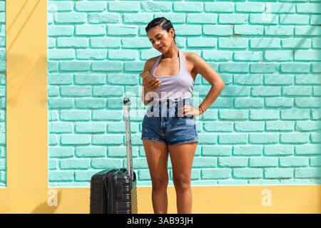 A young Latin woman stands against a colorful wall checking her phone next to luggage. The vibrant beach town atmosphere adds a lively touch. Stock Photo