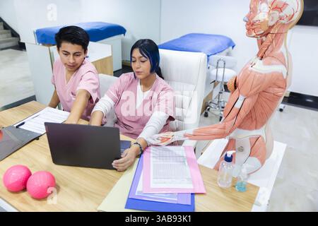 Two physiotherapists in pink scrubs collaborate using a laptop beside an anatomical model in a well-equipped clinic in Ecuador, emphasizing teamwork a Stock Photo