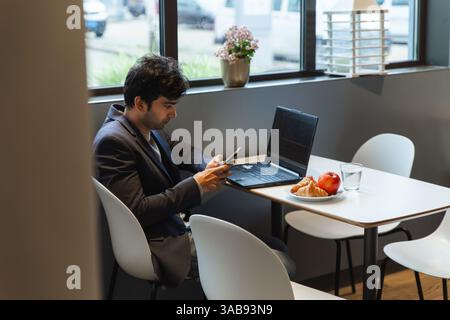 A indian man in a suit multitasks using a smartphone and laptop in a hotel cafeteria at a conference of AI in Amsterdam. A croissants, apple, and glas Stock Photo