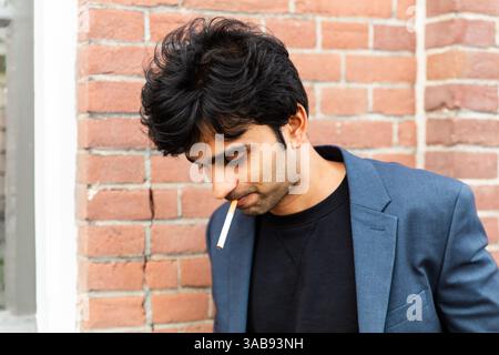 A indian man in a blue blazer stands against a brick wall, smoking. The casual pose and soft lighting add a contemplative mood to the scene during a c Stock Photo