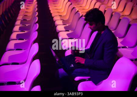 A indian man sits alone in an auditorium with purple lighting at a conference of AI in Amsterdam, working on a laptop and holding a coffee cup. Stock Photo