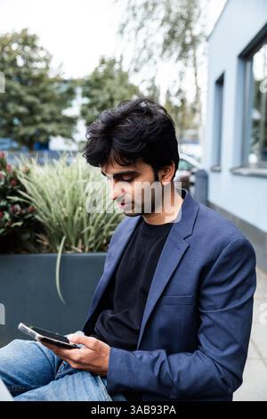 A indian man in a blue blazer and jeans is intently focused on his smartphone while sitting outdoors at a conference of AI in Amsterdam, surrounded by Stock Photo