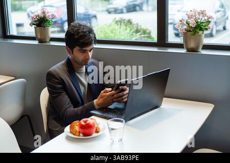 A indian man multitasks efficiently using a smartphone and laptop at a hotel cafe table in amsterdam during a conference of AI. He is enjoying a light Stock Photo