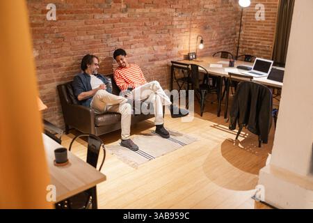 Two men sit on a sofa in a modern coworking space with brick walls and wooden floors. A laptop and table are in the background, fostering productivity Stock Photo
