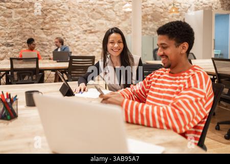 Professionals share a vibrant coworking space, collaborating and engaging in discussion They work on laptops at shared desks, reflecting teamwork and Stock Photo