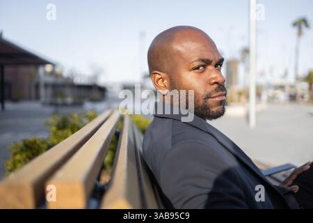 A cuban man in a suit sits on a bench in an urban environment, looking thoughtfully into the distance. The blurred background suggests a cityscape atm Stock Photo