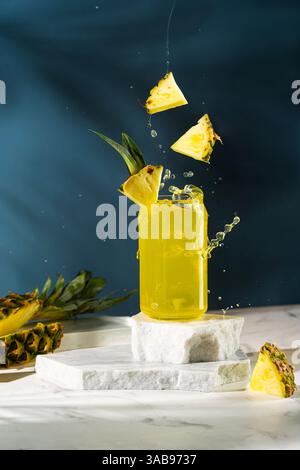 A vibrant pineapple drink in a glass jar with slices of pineapple splashing over it on a marbled platform against a dark blue background. Stock Photo