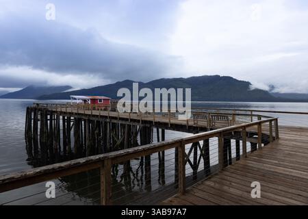 A serene landscape featuring a wooden pier extending into calm waters in Hoonah, Alaska, with a vibrant red building and a dramatic mountain range env Stock Photo