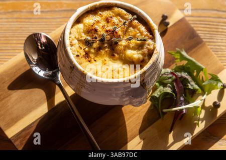 A sumptuous bowl of onion soup, topped with melted cheese and thyme, paired with a fresh side salad, presented on a rustic wooden board under natural Stock Photo