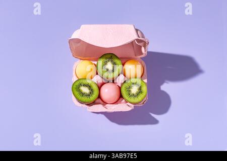 Top view of a pastel pink egg carton filled with colorful Easter eggs and freshly sliced kiwi fruits arranged creatively on a purple background. Stock Photo