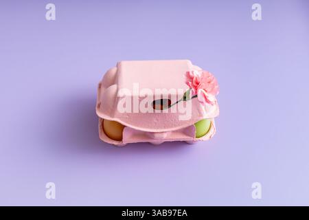 Top view of a pink egg carton decorated with a pink flower, containing colorful Easter eggs on a purple background. Stock Photo