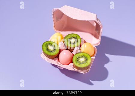 Top view of a pink egg carton holding slices of green kiwi and variously colored Easter eggs on a purple surface, showcasing a vibrant scene of fresh Stock Photo