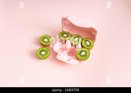 Top view of a creative arrangement of sliced kiwi fruits, resembling eggs, neatly placed in a pink egg carton on a light pink background. Stock Photo