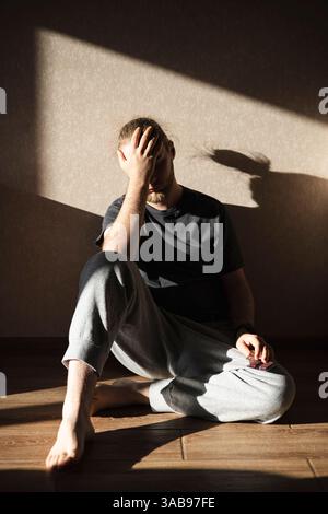 A man sits barefoot on the floor at home, deeply contemplating while holding a blister pack of pills. His expression conveys emotion, highlighting the Stock Photo