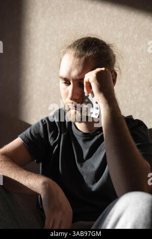 Young man with a beard thoughtfully holding a blister pack of pills. Sitting in a room, he appears deep in thought, suggesting themes of health or dec Stock Photo