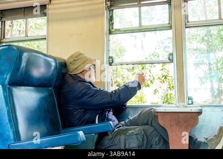 A caucasian elderly man in a blue jacket and beige hat sits in a train, looking out the window and taking picture with mobile phone at something inter Stock Photo