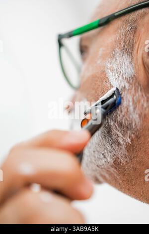 Close-up of a mature man shaving in the bathroom. The man, wearing glasses, uses a razor across a lathered face, showcasing a daily grooming routine. Stock Photo