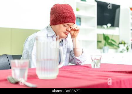 A man in a cozy red beanie and casual shirt sits at a table in a mental health residence, appearing thoughtful and reflective. A water jug and glass a Stock Photo