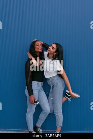 Two young friends stand against a blue wall, sharing headphones and enjoying music. They are smiling, expressing joy and friendship, dressed casually, Stock Photo
