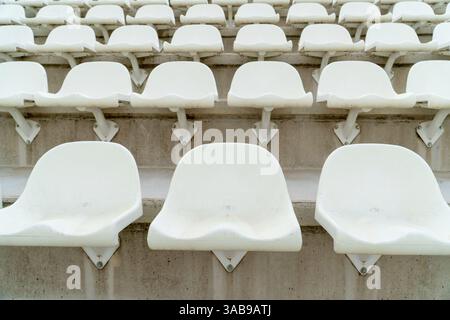 A section of empty, weathered white seats arranged in rows at an outdoor athletics stadium, emphasizing the quiet and stillness of an off-season or no Stock Photo