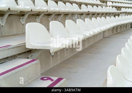 Rows of empty white seats await spectators in an outdoor athletics stadium, with visible seat numbers for organized seating. Stock Photo