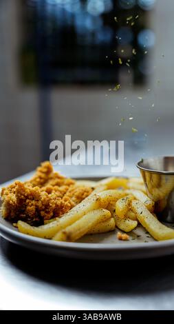 A close-up shot of crispy golden fries and crunchy chicken pieces on a plate. Fresh herbs are sprinkled above, creating a dynamic and appetizing cafe Stock Photo