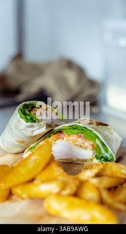 A close-up of a mouthwatering chicken wrap, filled with fresh lettuce and tomato, accompanied by crispy potato wedges on a wooden board. A perfect caf Stock Photo