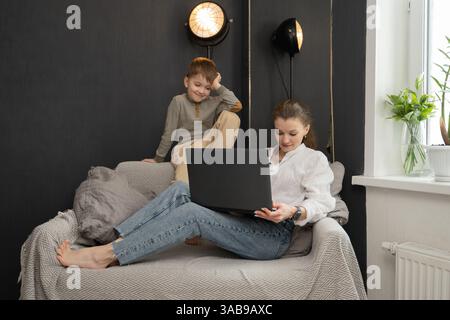 A mother lounges on a cozy sofa, working on her laptop, while her playful child sits nearby. The room features soft lighting and a decorative plant on Stock Photo