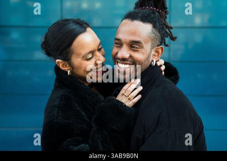 A happy multicultural couple embraces and smiles lovingly at each other, symbolizing joy and connection. They are wearing black coats against a modern Stock Photo