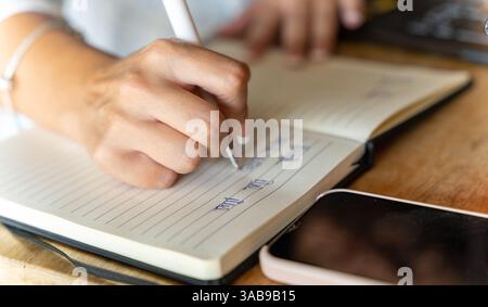 A woman writes in a lined notebook using a pen, with a smartphone placed beside them on a wooden table. This image captures a moment of focused note-t Stock Photo