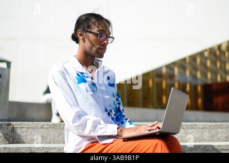 A thoughtful man wearing a floral shirt works on a laptop while seated outdoors. His attire and demeanor represent confidence, pride, and inclusivity Stock Photo