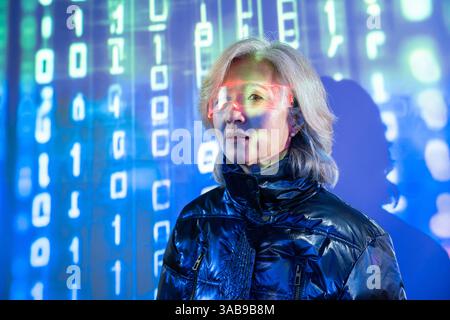 An elderly Asian woman is immersed in the metaverse, wearing futuristic eyewear. The digital background is filled with glowing binary code, symbolizin Stock Photo