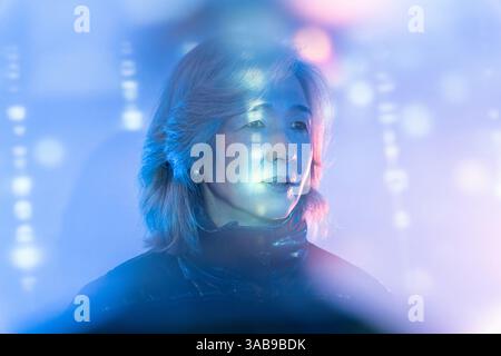 An elderly Asian woman interacts with a colorful metaverse interface, surrounded by ethereal blue and pink lights, showcasing technology's potential f Stock Photo