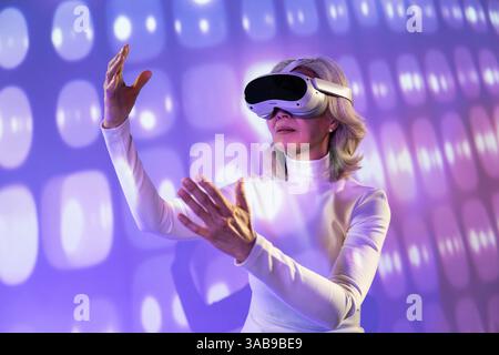 An elderly Asian woman is immersed in the metaverse, wearing virtual reality goggles. She stands in a futuristic, illuminated environment, interacting Stock Photo