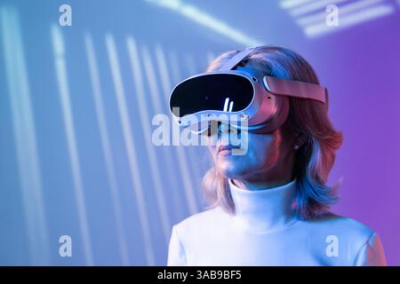 An elderly Asian woman wearing virtual reality goggles immerses herself in the metaverse. The image reflects the diversity of users engaging with adva Stock Photo