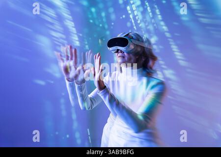 An elderly Asian woman engages with the metaverse using a virtual reality headset. She extends her hands to interact with digital elements, showcasing Stock Photo
